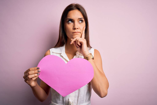jong mooi brunette vrouw met grote roze hart papier vieren valentijn dag serieus gezicht denken over vraag, zeer verward idee - Foto, afbeelding