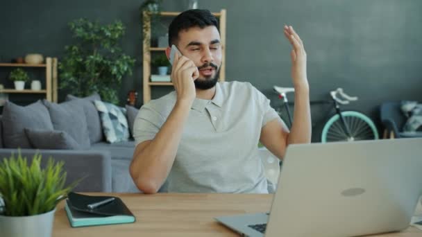 Emotional Arab man speaking on mobile phone and gesturing at table in apartment - Felvétel, videó