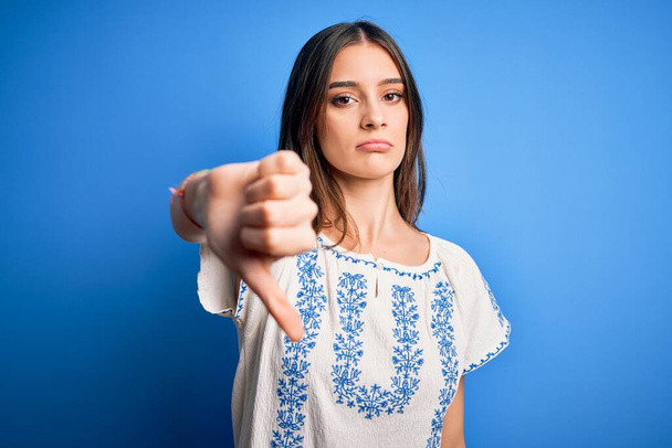 Nuori kaunis brunette nainen päällään rento t-paita seisoo sininen tausta näyttää onneton ja vihainen osoittaa hylkääminen ja negatiivinen peukalot alas ele. Huono ilme. - Valokuva, kuva