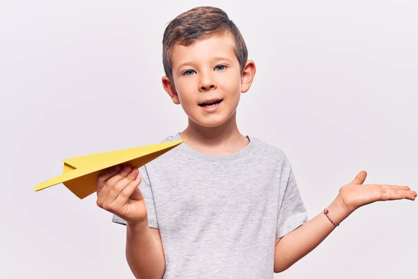 Miúdo loiro bonito segurando avião de papel celebrando a realização com sorriso feliz e expressão vencedora com mão levantada
  - Foto, Imagem