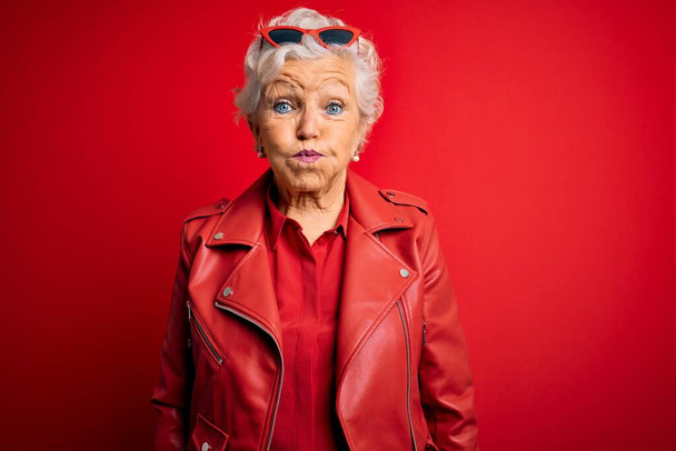 Senior mooie grijs-harige vrouw draagt casual rode jas en zonnebril puffen wangen met grappig gezicht. Mond opgeblazen met lucht, gekke uitdrukking. - Foto, afbeelding