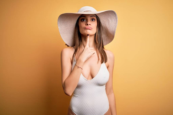 Młoda piękna brunetka na wakacjach ubrana w strój kąpielowy i letni kapelusz Myśląc o wątpliwości z palcem na podbródku i patrząc w górę zastanawiając - Zdjęcie, obraz