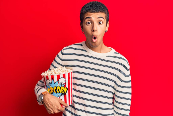 Junger afrikanisch-amerikanischer Mann hält Popcorn verängstigt und erstaunt mit offenem Mund für Überraschung, ungläubiges Gesicht  - Foto, Bild
