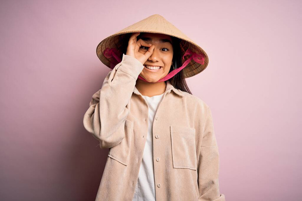 Junge schöne Frau, die traditionellen konischen asiatischen Hut über isoliertem rosa Hintergrund tut ok Geste mit der Hand lächelnd, Blick durch die Finger mit glücklichem Gesicht. - Foto, Bild