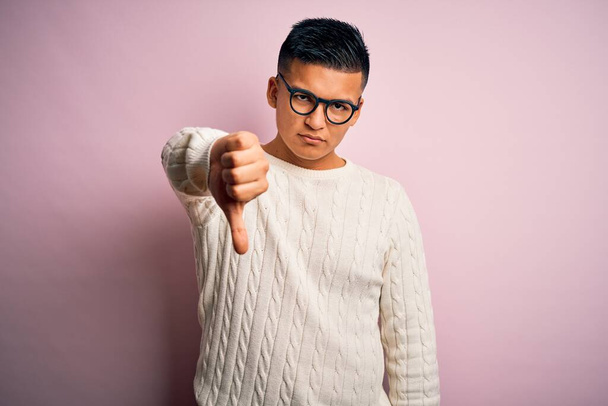 Homem latino bonito jovem vestindo suéter casual branco e óculos sobre fundo rosa olhando infeliz e irritado mostrando rejeição e negativo com polegares para baixo gesto. Má expressão
. - Foto, Imagem