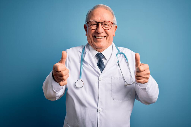 Älterer grauhaariger Arzt, der Stethoskop und Arztkittel über blauem Hintergrund trägt und eine positive Geste mit der Hand macht, die Daumen hoch lächelnd und glücklich. Fröhliche Miene und Siegergeste. - Foto, Bild