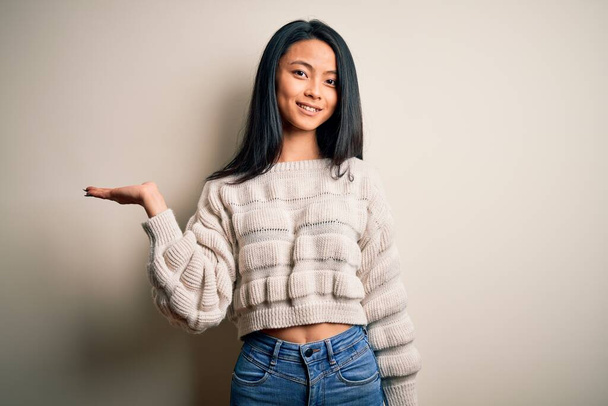 Young beautiful chinese woman wearing casual sweater over isolated white background smiling cheerful presenting and pointing with palm of hand looking at the camera. - Photo, Image