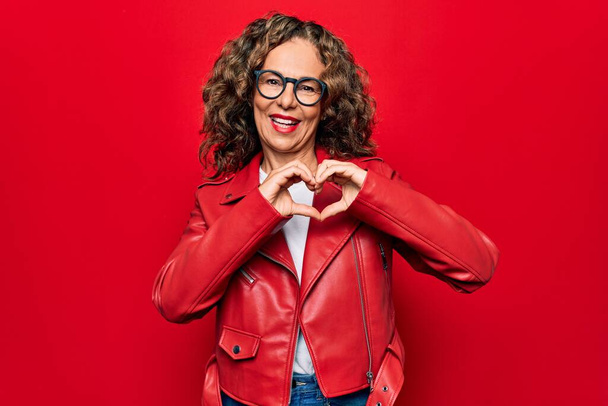Moyen Age belle femme portant une veste rouge décontractée et des lunettes sur fond isolé souriant en amour faisant forme de symbole de coeur avec les mains. Concept romantique. - Photo, image