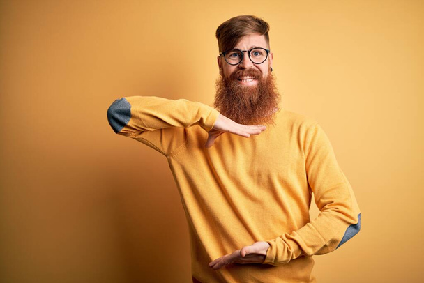 Handsome Irish redhead man with beard wearing glasses over yellow isolated background gesturing with hands showing big and large size sign, measure symbol. Smiling looking at the camera. Measuring concept. - Photo, Image