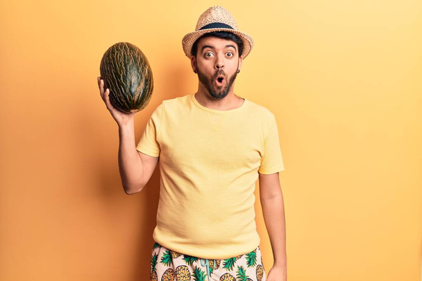 Junger hispanischer Mann in Badebekleidung und Sommermütze hält Melone erschrocken und erstaunt mit offenem Mund für Überraschung, ungläubiges Gesicht  - Foto, Bild