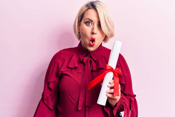 Beautiful blonde plus size woman holding graduated degree diploma over pink background scared and amazed with open mouth for surprise, disbelief face - Foto, afbeelding
