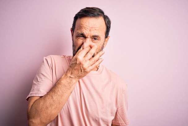 Mann mittleren Alters in lässigem T-Shirt vor isoliertem rosafarbenem Hintergrund, der etwas Stinkiges und ekelhaften, unerträglichen Geruch riecht und mit den Fingern auf der Nase den Atem anhält. Schlechter Geruch - Foto, Bild