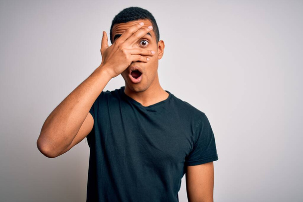 Young handsome african american man wearing casual t-shirt standing over white background peeking in shock covering face and eyes with hand, looking through fingers with embarrassed expression. - Fotoğraf, Görsel