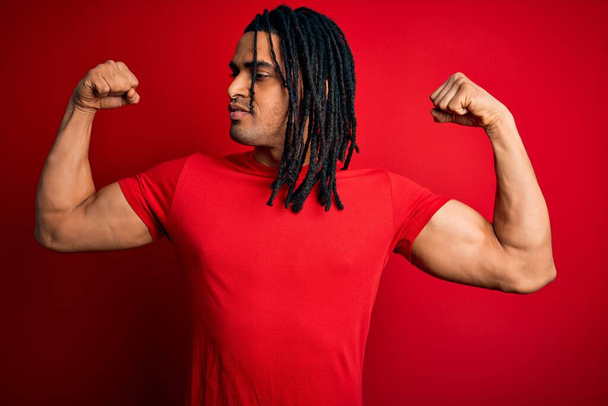 Kırmızı günlük tişörtlü, gururla gülümseyen kol kaslarını gösteren Afrikalı Afro Amerikalı genç bir adam. Fitness konsepti. - Fotoğraf, Görsel