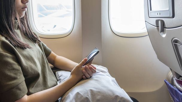 Τεχνολογία κατά την πτήση που ταξιδεύουν ή πετούν στο αεροπλάνο με νεαρό κορίτσι επιβάτη ή γυναίκα ταξιδιώτη χρησιμοποιώντας κινητό smartphone, online app και internet, ενώ κάθεται στο κάθισμα του αεροπλάνου κατά τη διάρκεια του ταξιδιού - Φωτογραφία, εικόνα