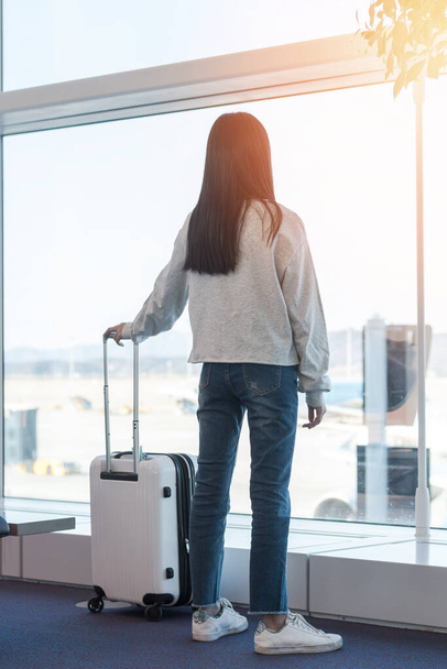 Lebensstil eines jungen Passagiers mit Gepäck in der Abflughalle des Flughafenterminals mit Blick auf das Flugzeug, das die Flugzeit und den Verspätungsstatus am Gate überprüft - Foto, Bild