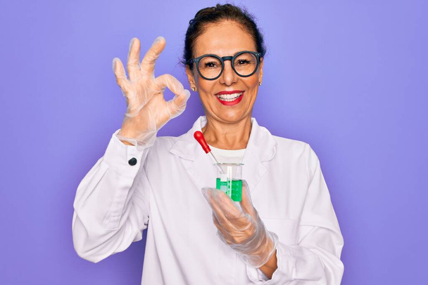 Старша вчена середнього віку, жінка в лабораторному пальто, що тримає дослідницьку пробірку, робить знак з пальцями, відмінний символ
 - Фото, зображення
