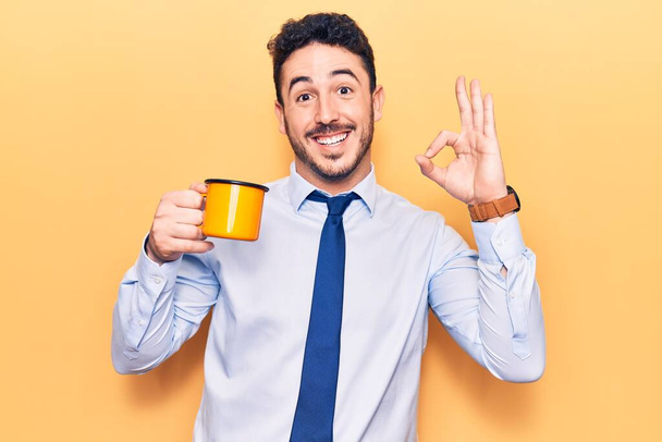 Young hispanic man wearing business clothes holding coffee doing ok sign with fingers, smiling friendly gesturing excellent symbol  - Φωτογραφία, εικόνα