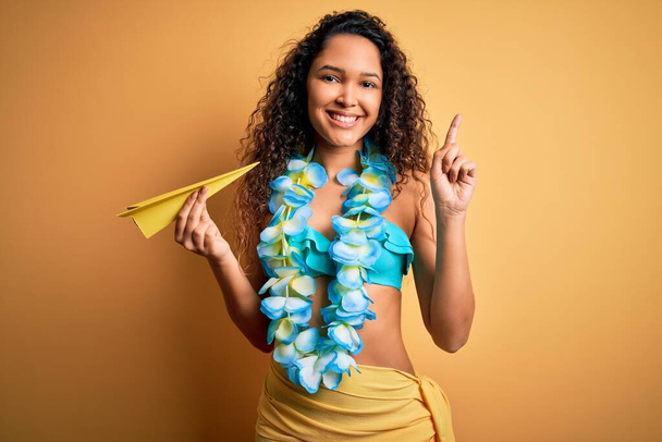 jonge mooie amerikaanse vrouw op vakantie dragen bikini holding papier vliegtuig verrast met een idee of vraag wijzend vinger met gelukkig gezicht, nummer een - Foto, afbeelding