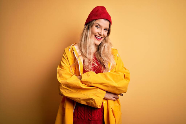 Jonge mooie blonde vrouw met regenjas en wollen pet over geïsoleerde gele achtergrond vrolijk gezicht glimlachend met gekruiste armen kijkend naar de camera. Positief persoon. - Foto, afbeelding