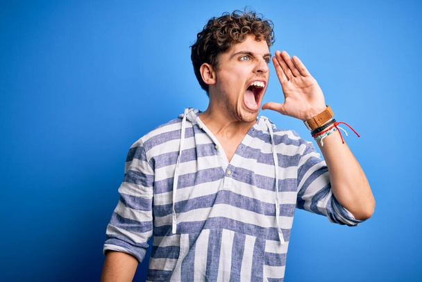 Young blond handsome man with curly hair wearing casual striped sweatshirt shouting and screaming loud to side with hand on mouth. Communication concept. - Photo, Image