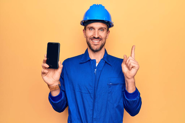 Fiatal jóképű munkás férfi visel egyenruhát és keménykalap gazdaság okostelefon mutatja képernyőn mosolygó boldog mutató kéz és ujj az oldalon - Fotó, kép