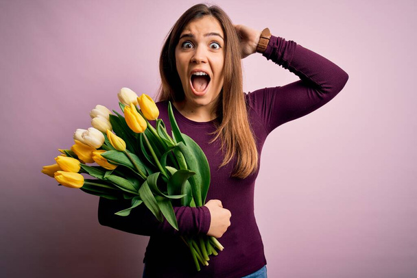 Young blonde woman holding romantic bouquet of yellow tulips flowers over pink background Crazy and scared with hands on head, afraid and surprised of shock with open mouth - Foto, Imagen