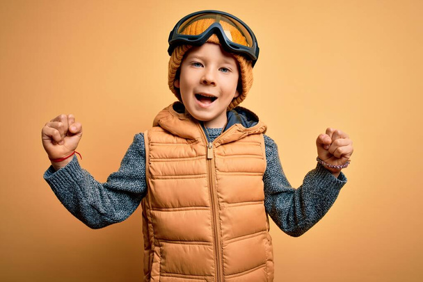 Young little caucasian kid wearing snow glasses and winter coat over yellow background celebrating surprised and amazed for success with arms raised and open eyes. Winner concept. - Foto, Bild