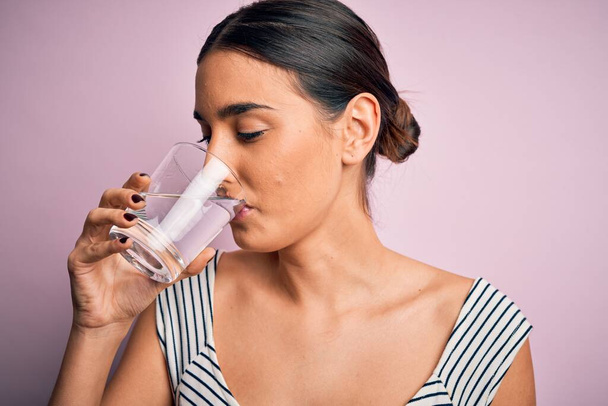 Jovem bela mulher morena bebendo copo de água saudável para refrescar em pé sobre fundo rosa isolado
 - Foto, Imagem