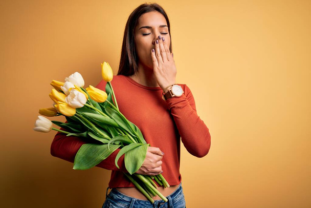 Jonge mooie brunette vrouw met boeket van tulpen bloemen over gele achtergrond verveelde gapen moe bedekken mond met de hand. Rusteloosheid en slaperigheid. - Foto, afbeelding