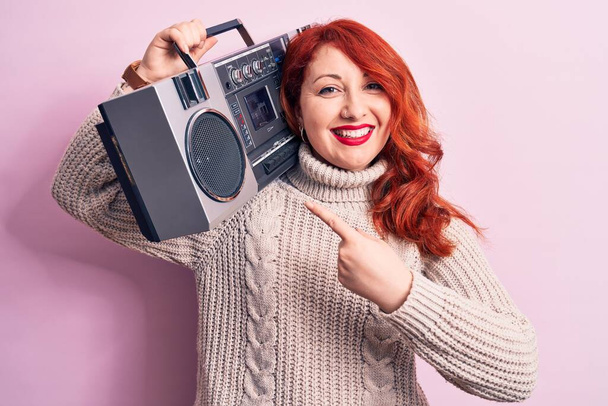 Kaunis punapää nainen kuuntelee musiikkia tilalla vintage boombox vaaleanpunainen tausta hymyillen onnellinen osoittaa käsin ja sormella - Valokuva, kuva