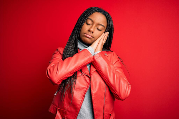 Молода афроамериканська жінка, одягнена в прохолодну моду шкіряну куртку над червоним ізольованим фоном, спить втомлений сон і складає руки разом, посміхаючись закритими очима. - Фото, зображення