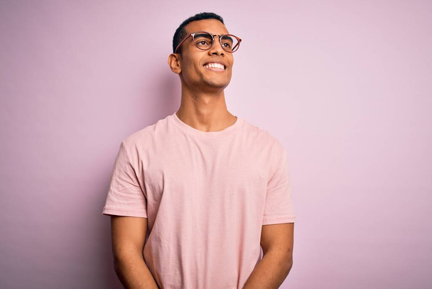Hombre afroamericano guapo vistiendo camiseta casual y gafas sobre fondo rosa mirando hacia un lado con sonrisa en la cara, expresión natural. Riendo confiado
. - Foto, imagen