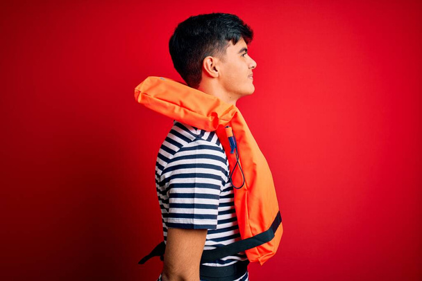 Young handsome man wearing orange safety life jacket over isolated red background looking to side, relax profile pose with natural face with confident smile. - Photo, image