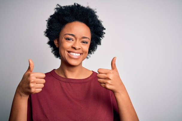 Mladá krásná afroamerická afro žena s kudrnatými vlasy na sobě ležérní tričko stojící úspěch znamení dělá pozitivní gesto s rukou, palce nahoru s úsměvem a šťastný. Veselý výraz a gesto vítěze. - Fotografie, Obrázek