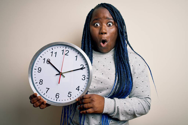 Afrika kökenli Amerikalı, saçları örgülü, elinde büyük bir saat tutan, şaşırmış ve korku ifadesinden korkan genç bir kadın. - Fotoğraf, Görsel