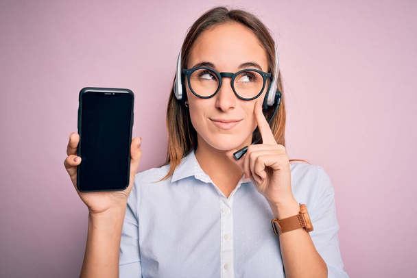Beautiful call center agent woman working using headset holding smartphone holding screen serious face thinking about question, very confused idea - Foto, afbeelding