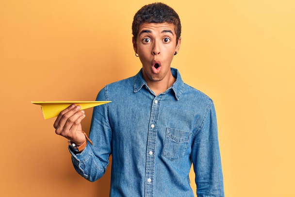 Joven africano amerciano sosteniendo avión de papel asustado y sorprendido con la boca abierta para sorpresa, cara de incredulidad
  - Foto, Imagen
