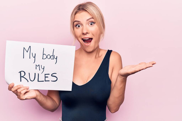 Young blonde woman asking for women freedom holding paper with my body my rules message celebrating achievement with happy smile and winner expression with raised hand - Photo, Image