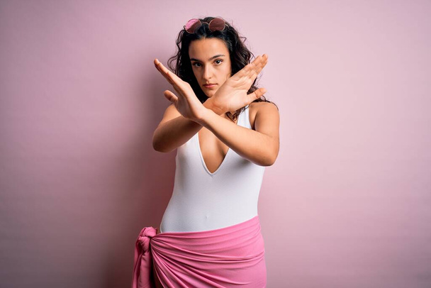 Mulher bonita com cabelos cacheados em férias vestindo maiô branco sobre fundo rosa Expressão de rejeição cruzando braços fazendo sinal negativo, rosto zangado
 - Foto, Imagem