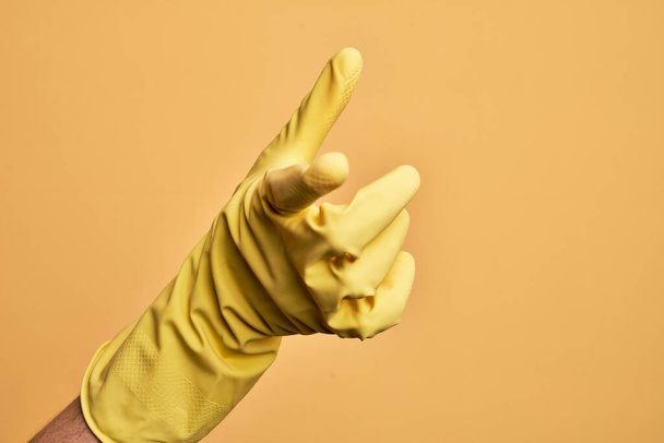 Mão de jovem caucasiano com luva de limpeza sobre fundo amarelo isolado apontando indicador para a câmera, escolhendo e indicando para a direção
 - Foto, Imagem