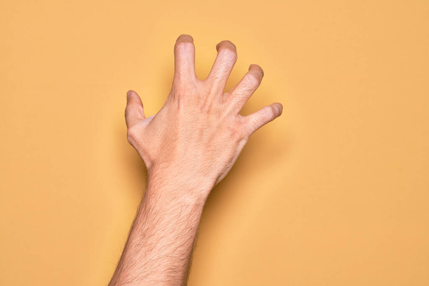 Hand van Kaukasische jonge man met vingers over geïsoleerde gele achtergrond grijpen agressief en eng met vingers, geweld en frustratie - Foto, afbeelding