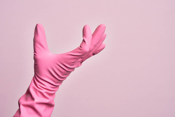 Ręka białego młodzieńca z rękawicą czyszczącą nad odizolowanym różowym tłem zbierającym i zabierającym niewidzialną rzecz, trzymającym obiekt z palcami pokazującymi przestrzeń - Zdjęcie, obraz
