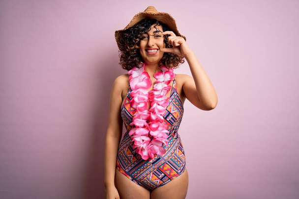 Joven mujer árabe hermosa de vacaciones con traje de baño y flores de lei hawaiano sonriendo y gesto seguro con la mano haciendo letrero de tamaño pequeño con los dedos mirando y la cámara. Concepto de medición
. - Foto, Imagen