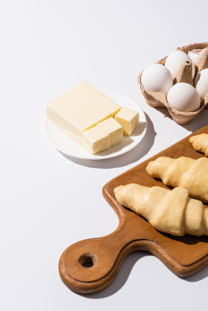 raw croissants on wooden cutting board near butter, eggs on white background - Fotografie, Obrázek