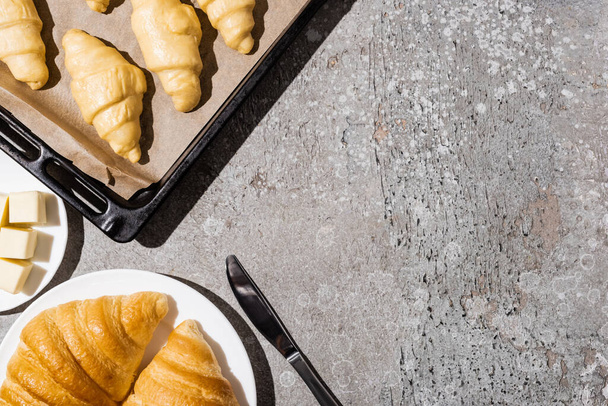 bovenaanzicht van ongekookte croissants op bakplaat nabij gekookt op plaat en boter op betonnen grijs oppervlak - Foto, afbeelding