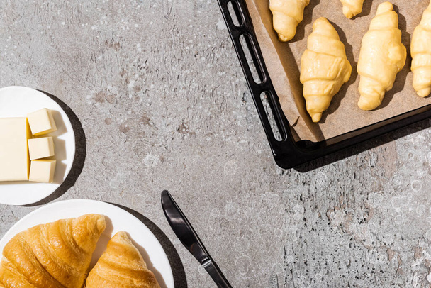 vista superior de croissants sin cocer en bandeja para hornear cerca de cocinado en plato y mantequilla en superficie gris hormigón
 - Foto, Imagen