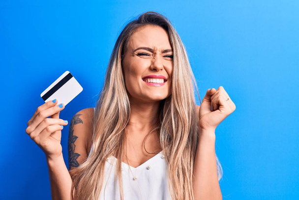 Young beautiful blonde woman holding credit card over isolated blue background screaming proud, celebrating victory and success very excited with raised arm - Photo, Image