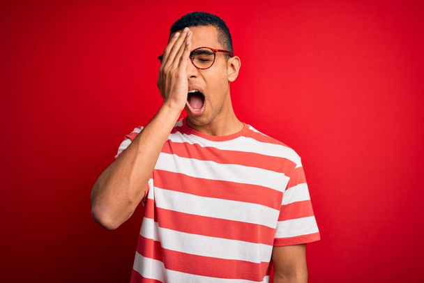 Mladý pohledný afroameričan v neformálním pruhovaném tričku a brýlích Zívání unaveně zakrývající půlku obličeje, oko a ústa rukou. Tvář bolí bolestí. - Fotografie, Obrázek