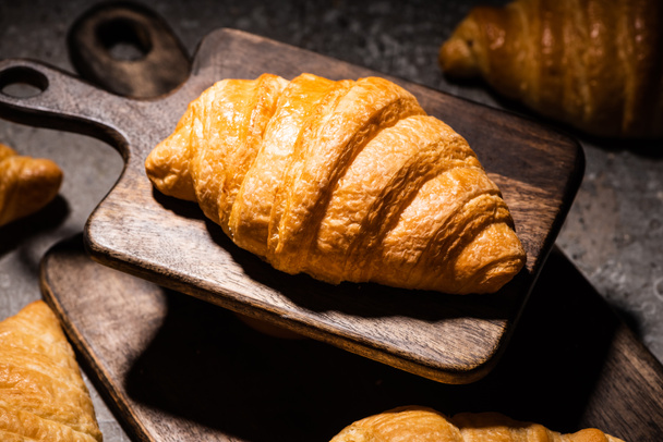 selective focus of fresh baked croissant on wooden cutting board on concrete grey surface in dark - Φωτογραφία, εικόνα